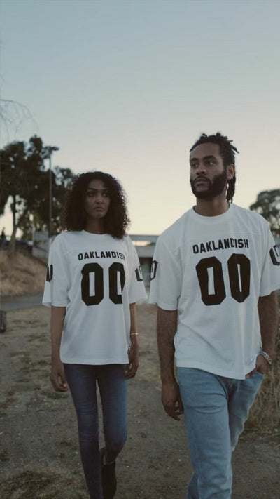 A woman and man standing outside in mesh Oaklandish football jerseys. 