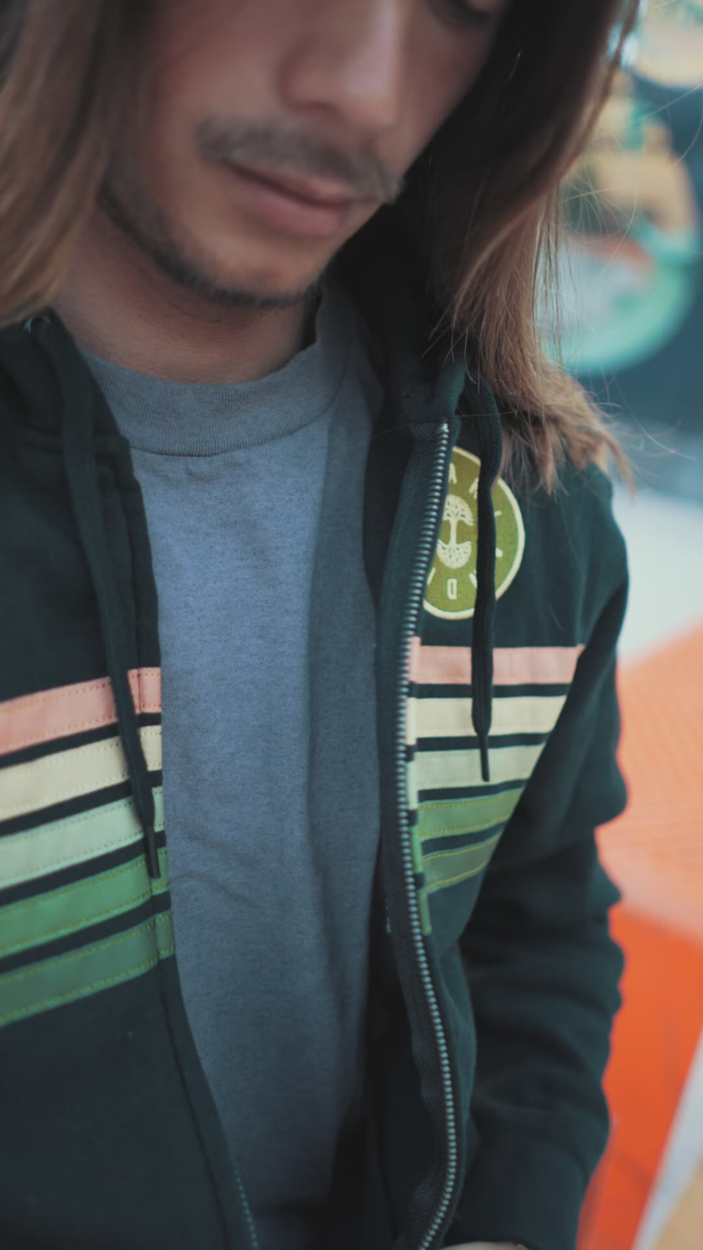 Male model with long hair in Downtown Oakland wearing black zip hoodie with stripes that he zips up and down, smiling looking away.