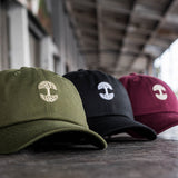 Three cotton dad caps in olive green, burgundy, and black sitting outdoors in a line.
