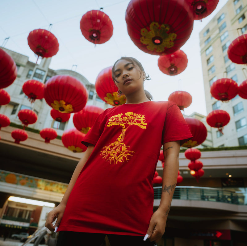 Female model wearing red t-shirt with gold dragon power graphic design in the shape of an Oaklandish tree logo while standing under lanterns.