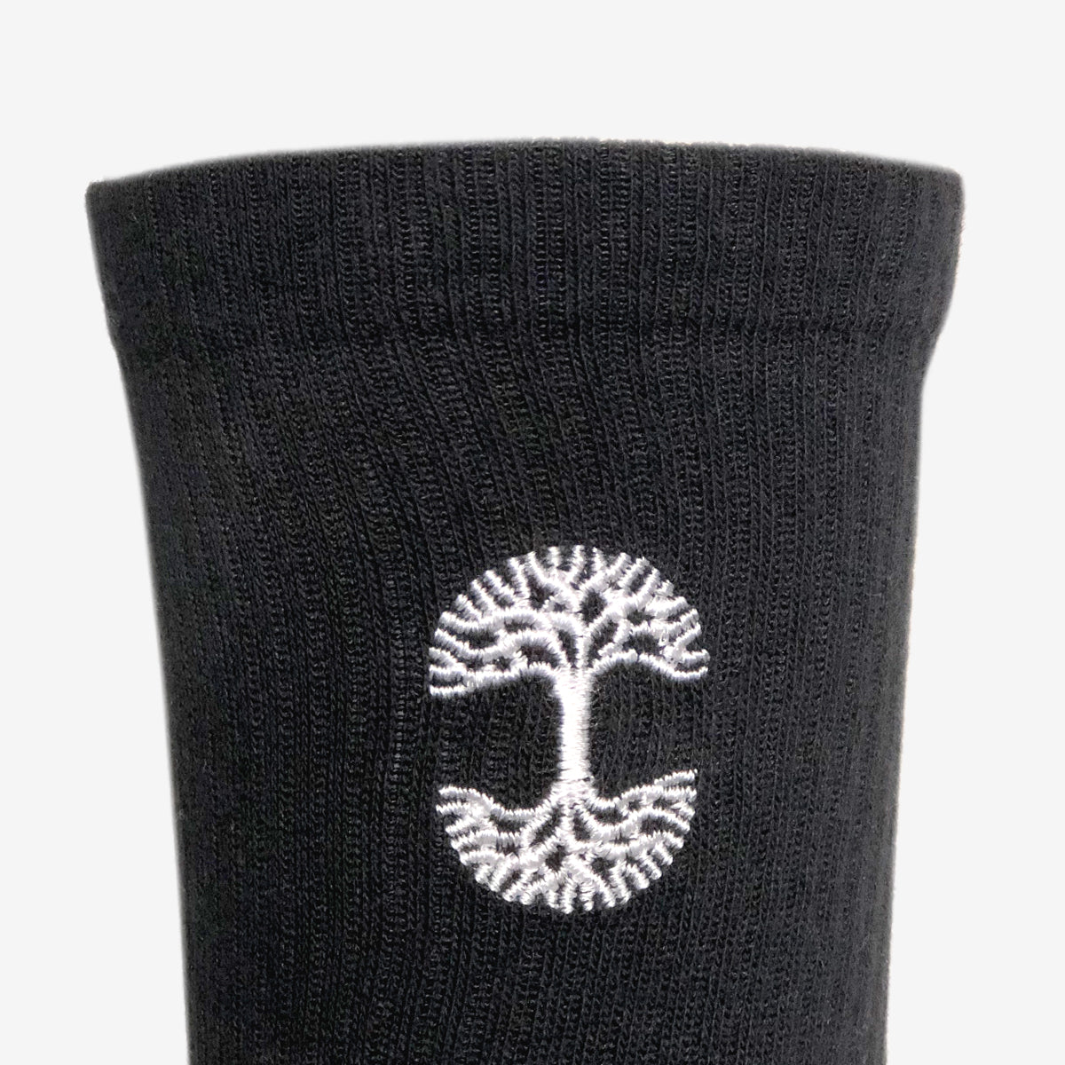 Close-up image of a pair of Oaklandish Embroidered Logo Socks in adult sizes, featuring a white embroidered design near the top. The design showcases a tree with roots in a circular formation, symbolizing growth and stability. The texture of the ribbed knit fabric is clearly visible against a plain white background.