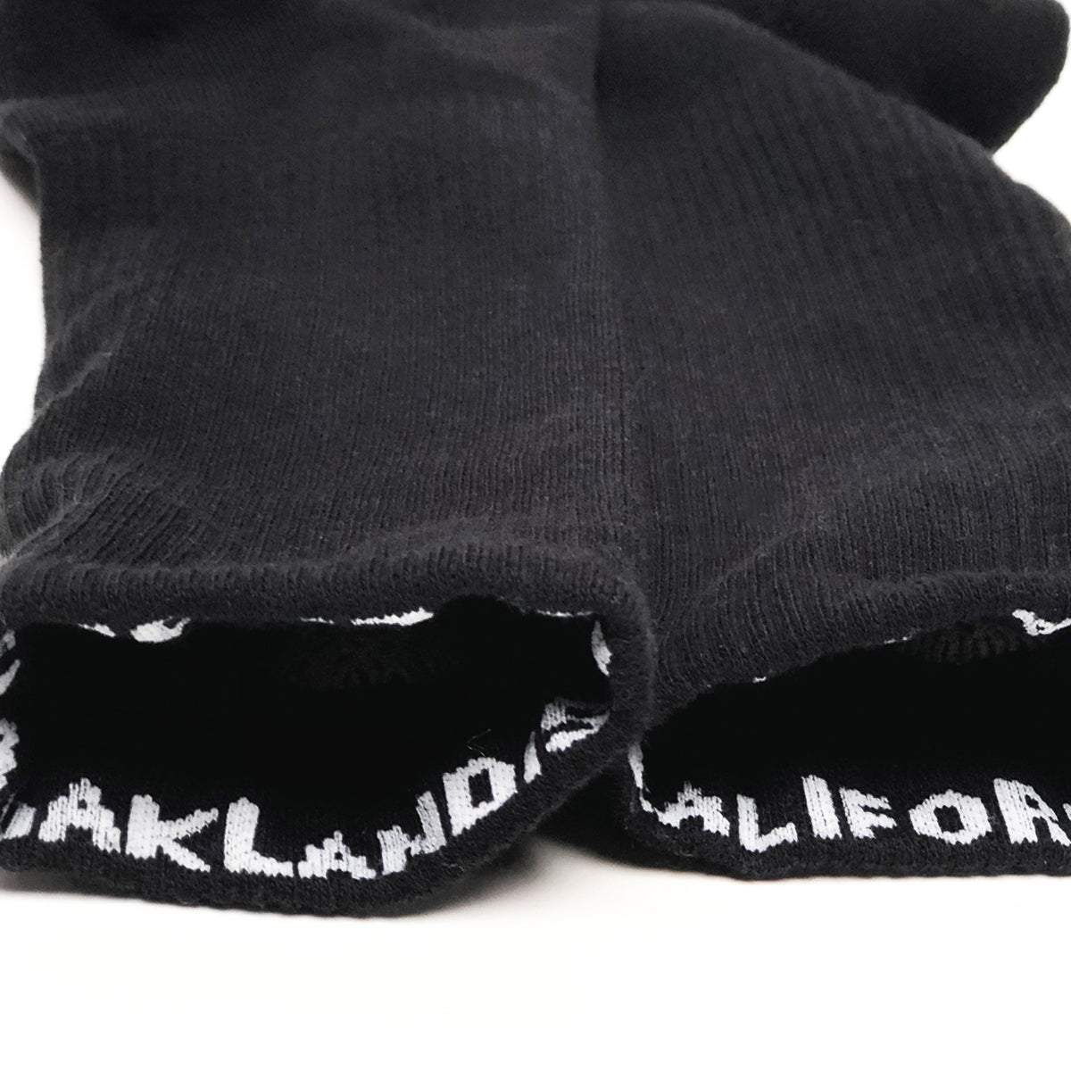 A pair of black Embroidered Logo Socks from Oaklandish is laid flat on a white surface. The cuff of each sock features the text "OAKLAND CALIFORNIA" in white, capital letters. The socks' fabric texture and knitted structure are visible, designed for adult sizes with a stylish nod to the brand's signature aesthetic.