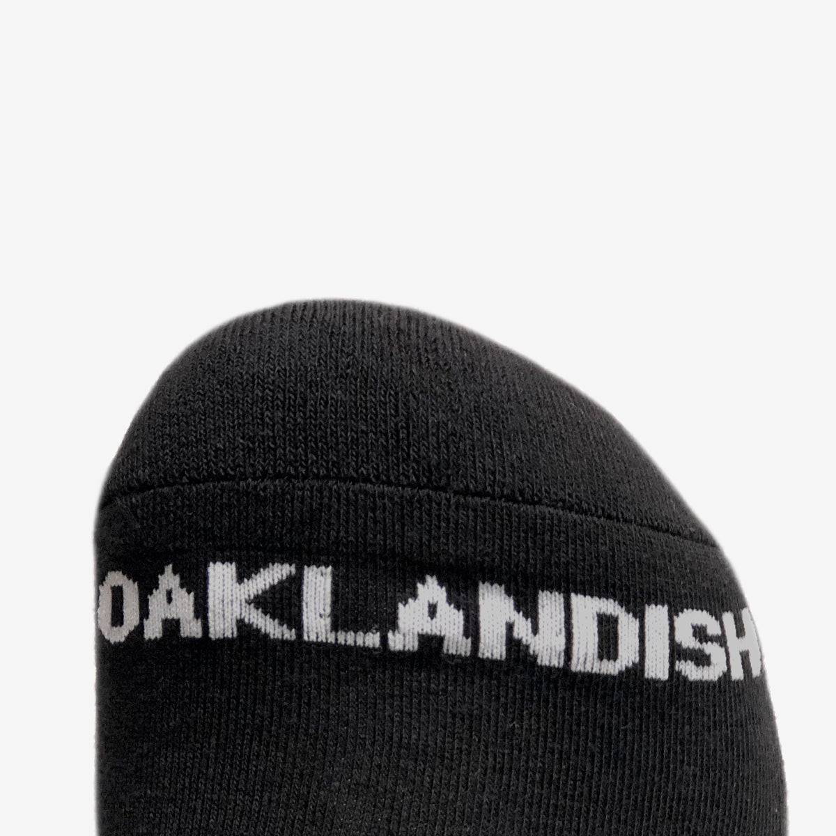 Close-up of the toe section of a black crew sock from the Oaklandish brand's Embroidered Logo Socks collection. The sock features "OAKLANDISH" in white, capital letters, prominently displayed near the front. The background is plain and light-colored, offering high contrast to the black sock and white text. These socks are available in adult sizes.