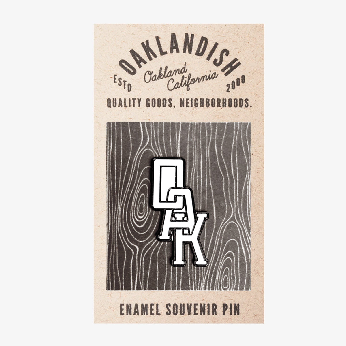 A rectangular card showcases the Enamel Pin - OAK Monogram featuring the letters "OAK" arranged vertically. The wood grain background displays text reading "Oaklandish," "Oakland California," "Est 2000," and "Quality Goods, Neighborhoods." The sleek white and black colors add a unique touch to this accessory from Oaklandish.
