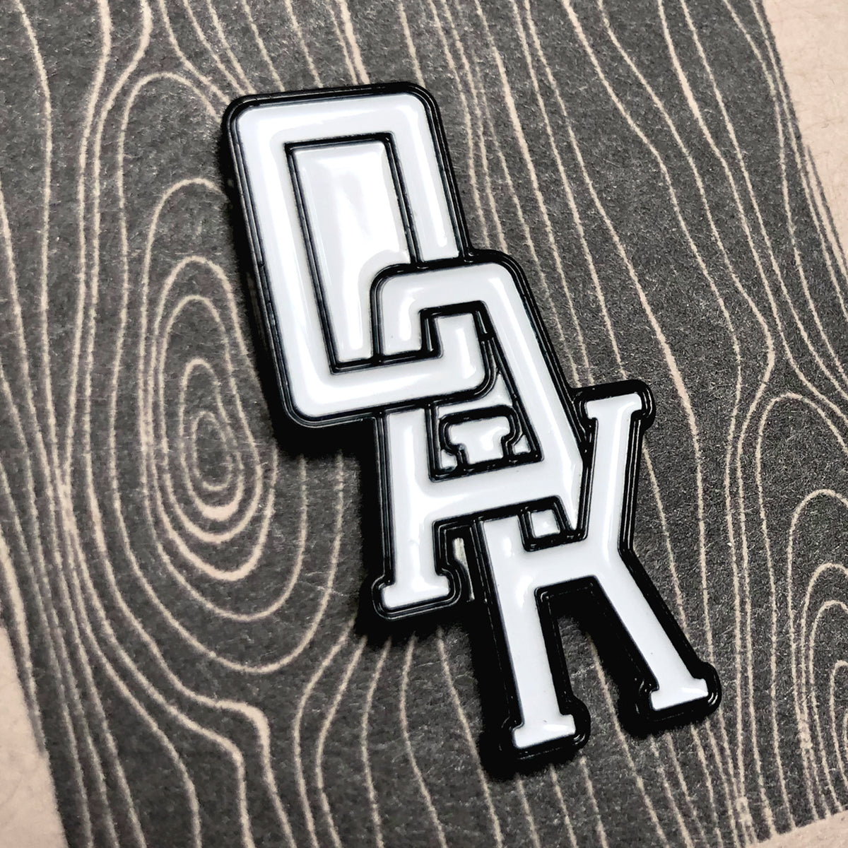 An Enamel Pin - OAK Monogram from Oaklandish lies on a textured surface featuring a wood grain pattern. The black and white pin showcases the letters "OAK" in a stylized, interlocking design, with the "O" and "A" positioned atop the "K." The bold, outlined letters make it a standout accessory.
