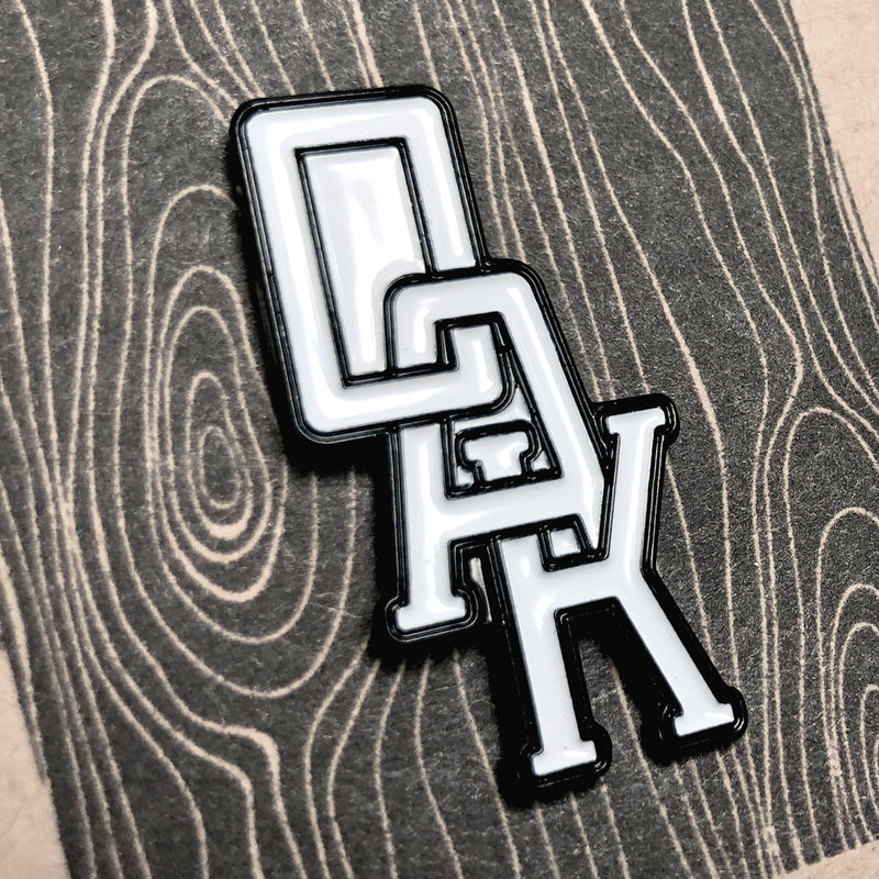 Close-up of white OAK wordmark with black trim on an enamel lapel pin.