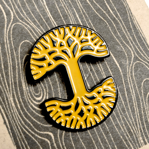Close up of enamel pin with yellow Oaklandish tree logo with black trim on brown paper retail packaging. 