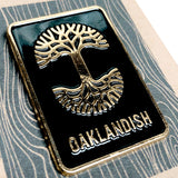 Close-up of enamel pin with gold Oaklandish tree logo and wordmark on black background on brown paper retail packaging. 