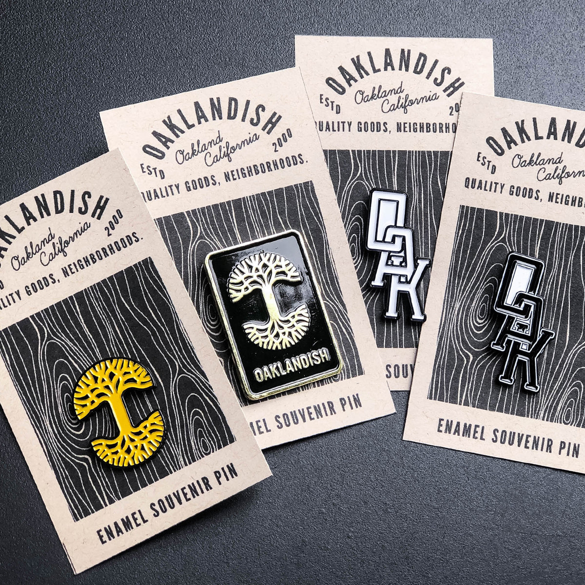 Four "Oaklandish" enamel lapel pins are displayed on rectangular beige backing cards with stylized black text. Two accessories feature the yellow and white Oaklandish tree logo, while the other two showcase the "OAK" monogram in bold lettering. The text on each card reads, "Oaklandish, Oakland California, Estd. 2000, Quality Goods, Neighborhoods.