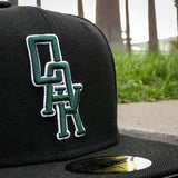 Close-up of green and white embroidered OAK wordmark on the crown of a black New Era cap on a sidewalk.