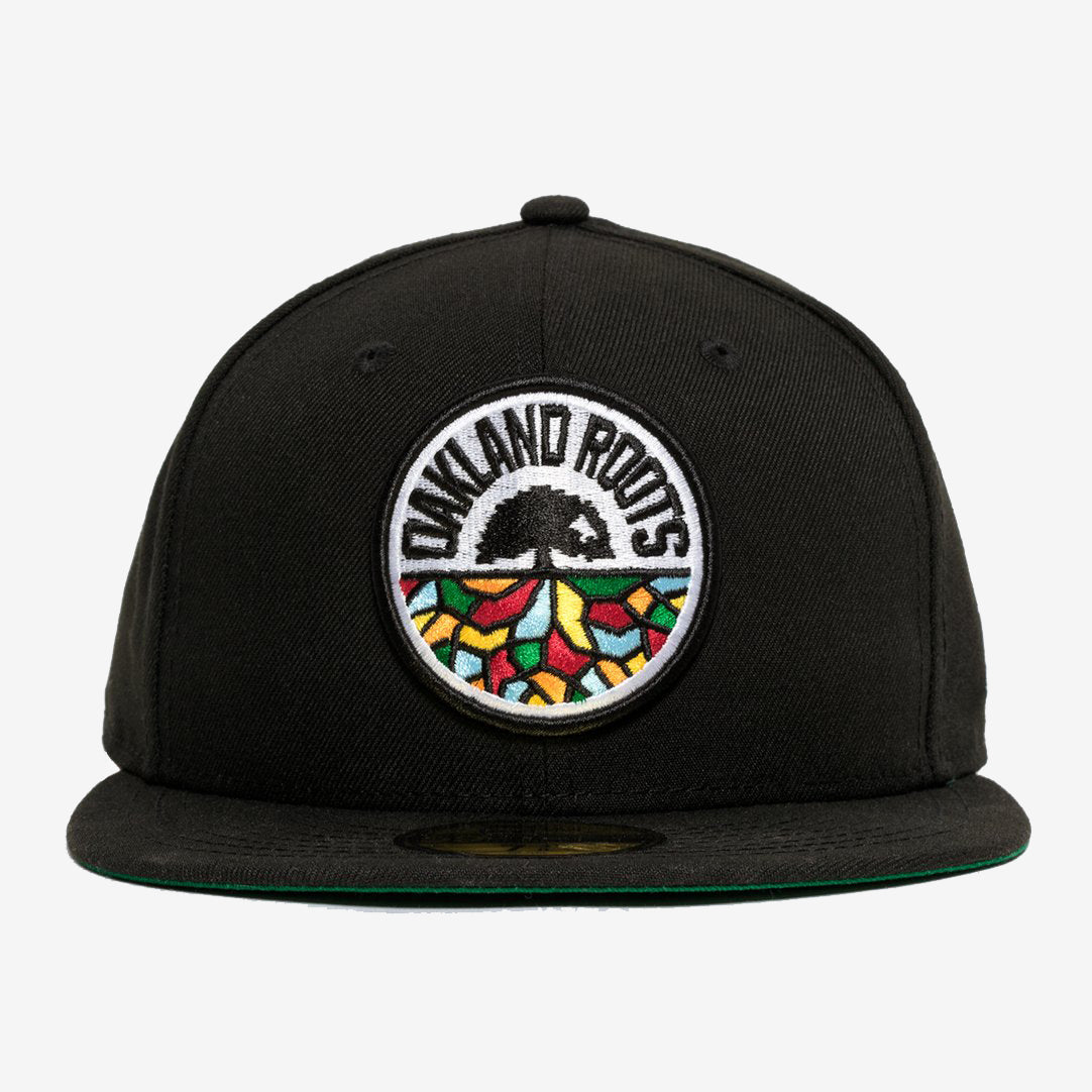 A New Era Oakland Roots SC 59FIFTY Fitted Cap in black, featuring a circular embroidered patch on the front. The patch showcases the words "Oakland Roots" at the top, a black tree in the center, and colorful geometric shapes resembling a mosaic at the bottom. This sports club cap comes with a flat brim and a green underside.