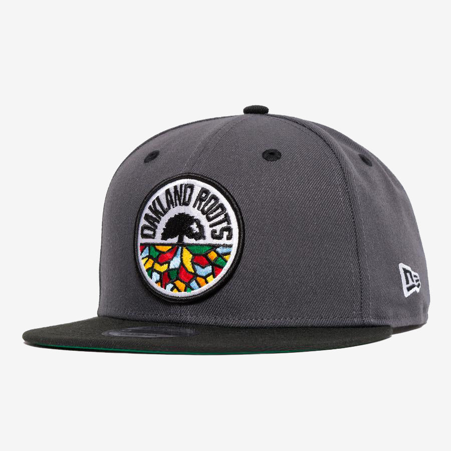 The New Era Oakland Roots SC 59FIFTY Fitted cap, in dark gray, showcases the Oakland Roots Sports Club logo with vibrant embroidery on the front panel. The logo features a tree with roots spreading out in various colors. The New Era logo is stitched onto the side, and the underside of the brim is dark green.