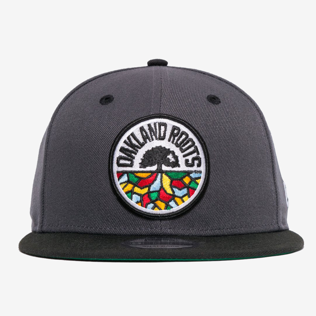 The New Era Oakland Roots SC 59FIFTY Fitted cap, in dark grey, showcases an intricately embroidered logo on the front featuring a black tree with spreading roots. Surrounding the tree is the bold text "OAKLAND ROOTS," accented by colorful shapes in red, yellow, green, and blue that symbolize the foundational pride of this distinguished sports club.