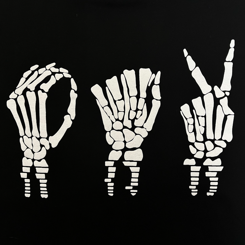  Close-up of skeletal bone hands signing 'OAK' in white puff ink on a black t-shirt.