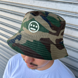 Man standing outdoors, wearing a Woodland Camo bucket hat with a Hieroglyphics Hip Hop Crew logo on the front crown. 