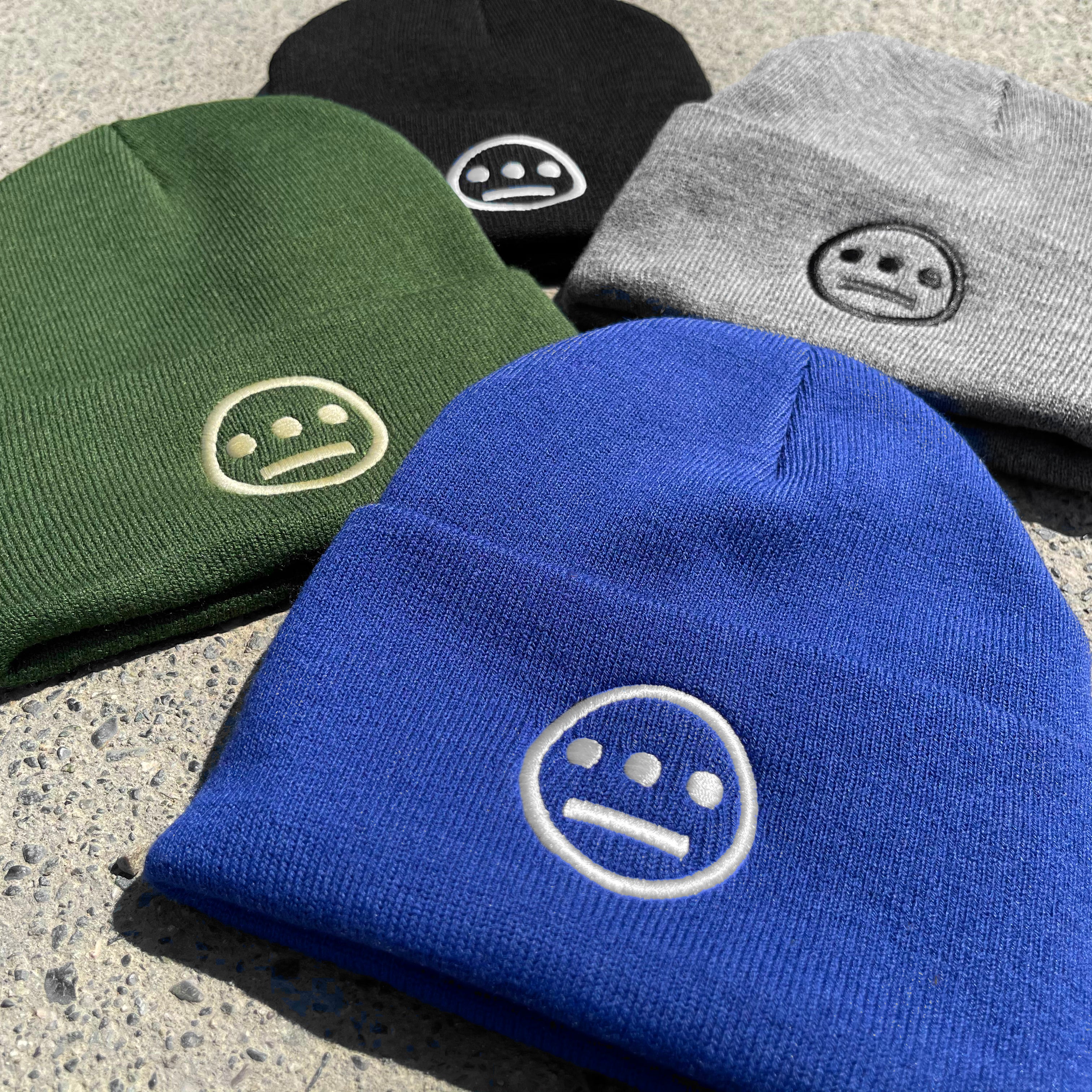 Close-up of 4 cuffed acrylic beanies in green, blue, black & grey, with white embroidered Hiero logos on on ashpalt.