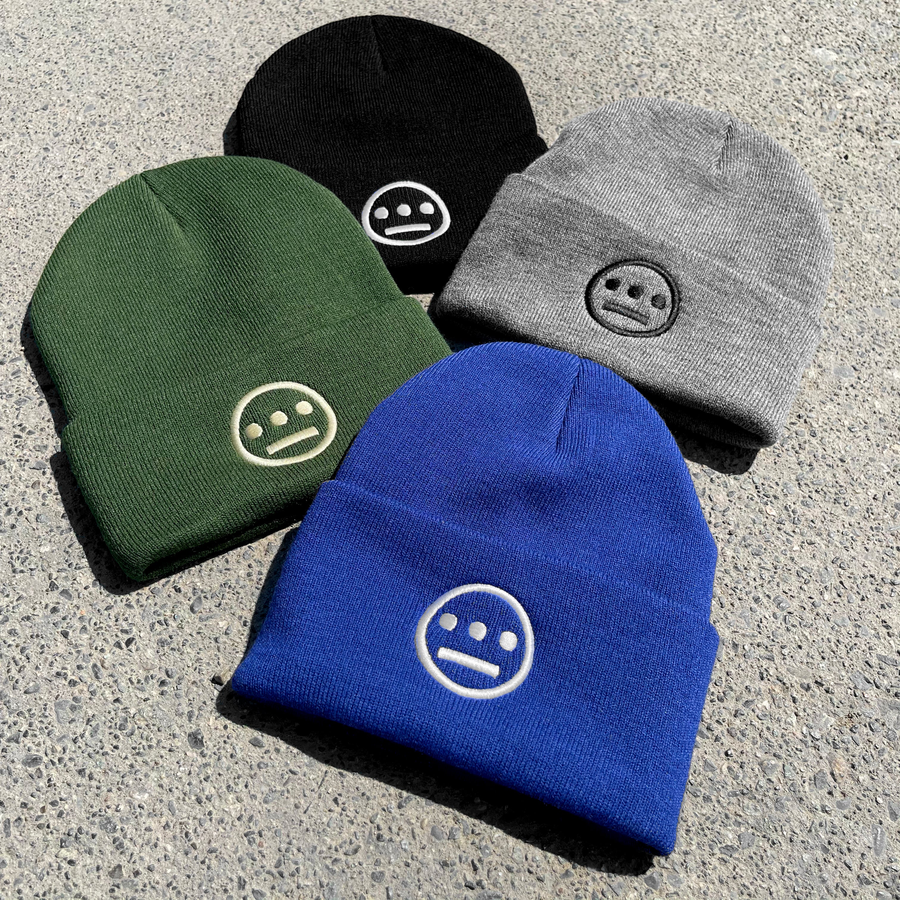The textured surface displays four Hiero Classic Cuff Beanies by Hieroglyphics, available in black, grey, green, and blue. Each beanie features an embroidered minimalist smiley face with eyes resembling hieroglyphics—a horizontal line with two dots—consistent across all the hats.