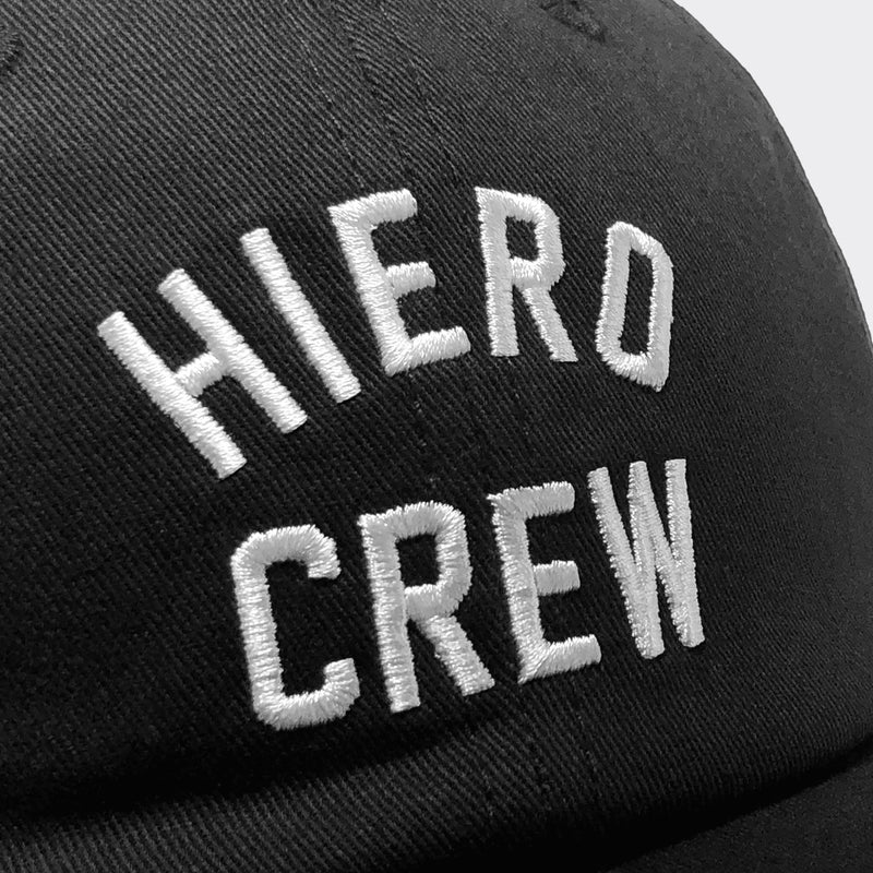 Close-up of white embroidered “HIERO CREW” logo on the crown of a black dad cap.