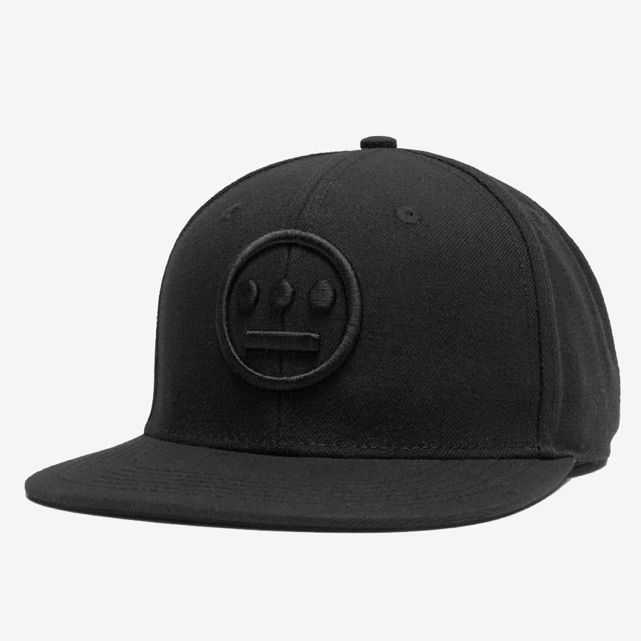 A black Hiero Logo Snapback by Hieroglyphics is displayed. The adjustable snapback cap features an embroidered, tone-on-tone circular emblem with three dots above a straight line, resembling a minimalist face, positioned on the front panel. The background is white, highlighting the hip hop-inspired design.
