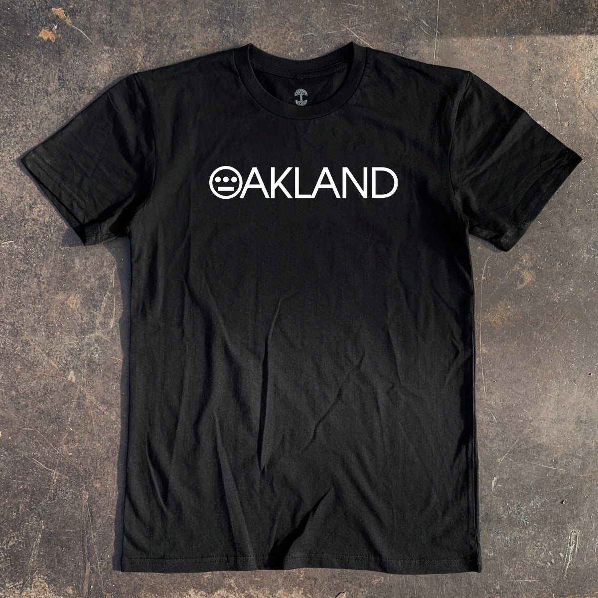 Black t-shirt with white OAKLAND wordmark with Hieroglyphics hip-hop crew logo as the O laying on asphalt.