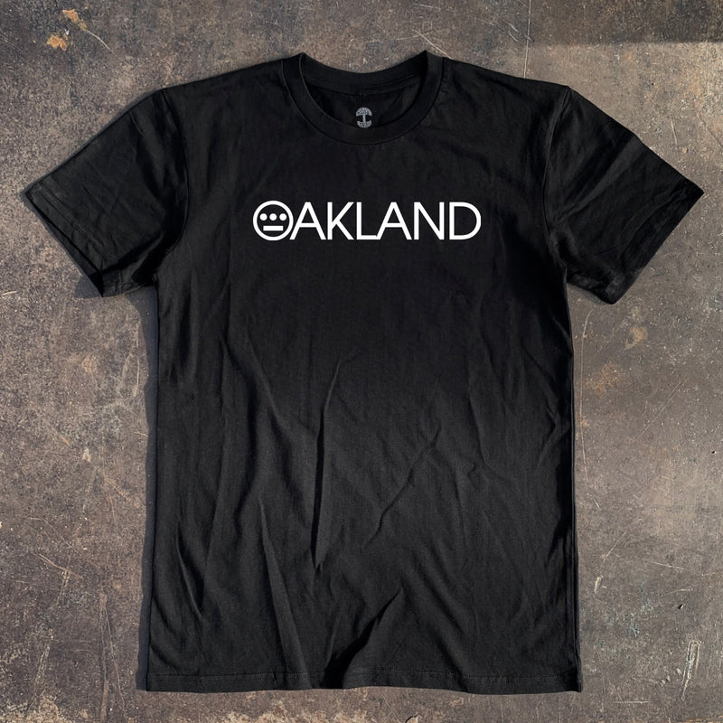 Black t-shirt with white OAKLAND wordmark with Hieroglyphics hip-hop crew logo as the O laying on asphalt.