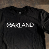 Close-up of white OAKLAND wordmark with Hieroglyphics hip-hop crew logo as the O on a black t-shirt laying on asphalt.