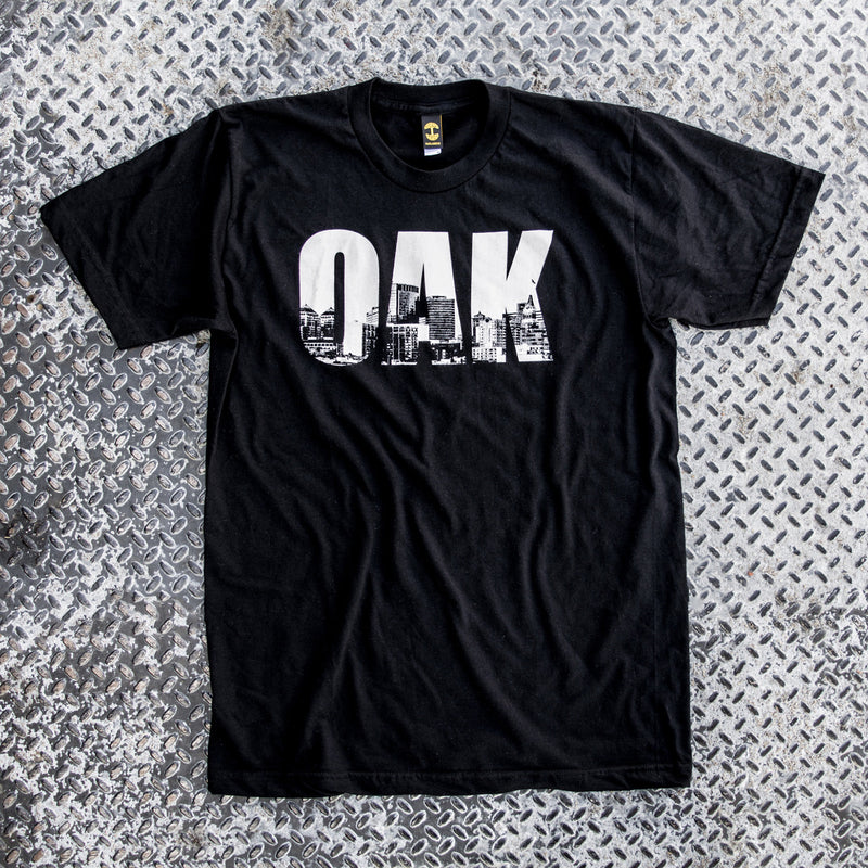 Black t-shirt with white capital letters spelling OAK, containing pictures of the Oakland skyline on grey background. 