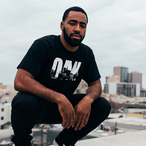 Black t-shirt with white capital letters spelling OAK, containing pictures of the Oakland skyline on male model. 