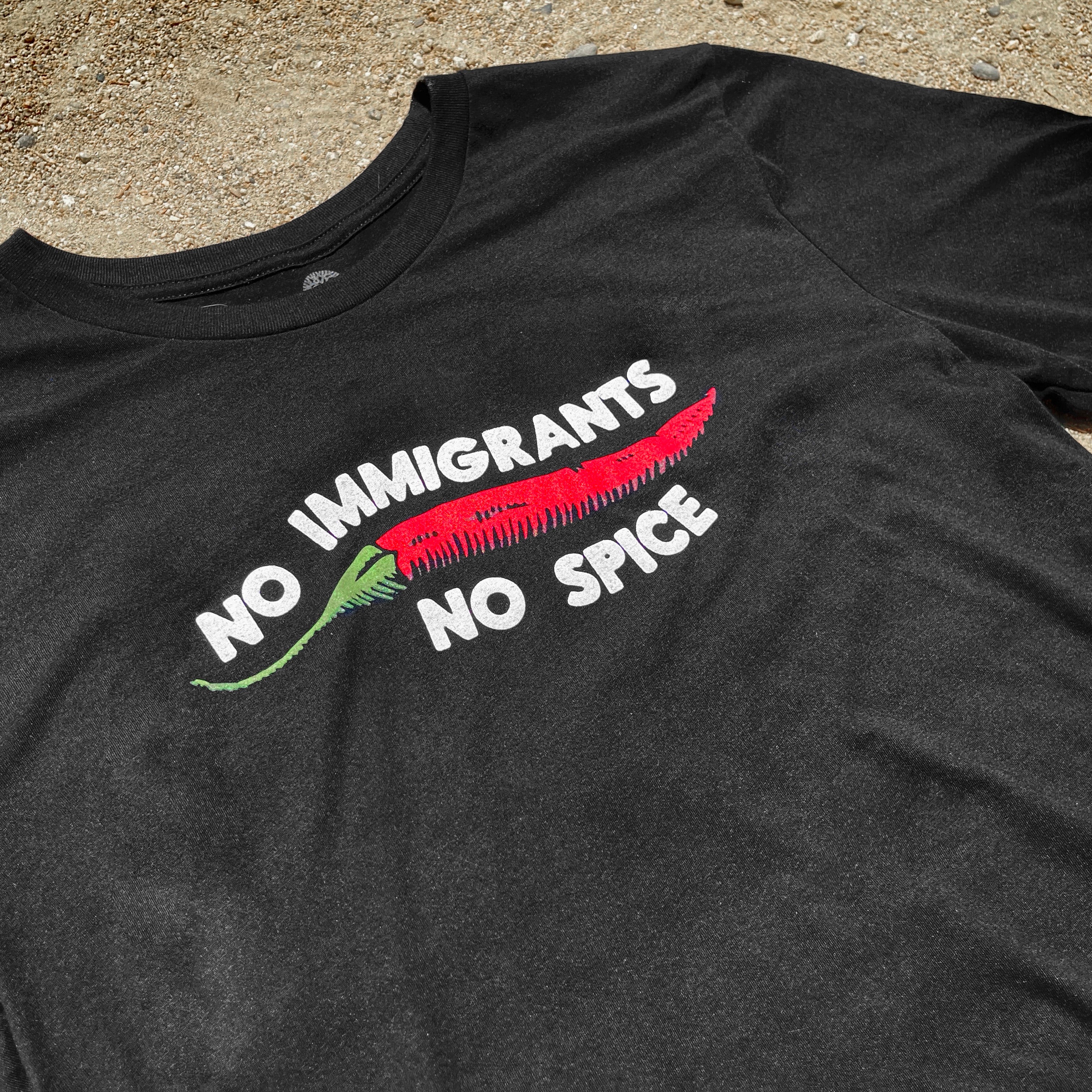 Close-up of a black t-shirt with No Immigrants, No Spice wordmark, and red chili pepper laying on the sand.