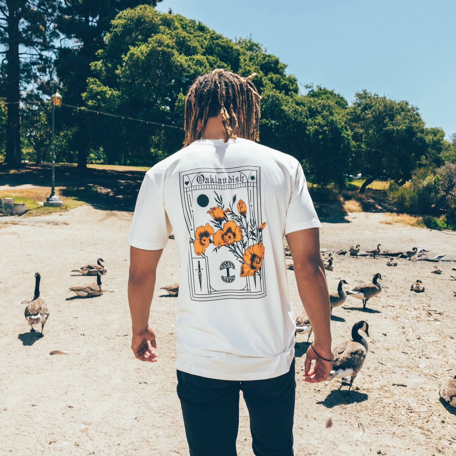 A man walking on the beach with geese showing the back side of a natural-color Blossom Oaklandish cotton t-shirt. 