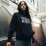 Women standing outdoors in Oakland wearing a black hooded sweatshirt with OAK in capital letters with a photo of Oakland cranes in each letter.