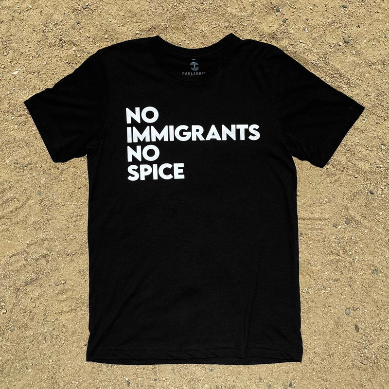 Black t-shirt with white No Immigrants, No Spice wordmark laying on sand.