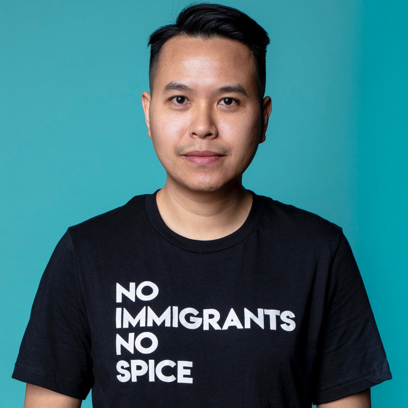 Man wearing a black t-shirt with white No Immigrants, No Spice wordmark on it with a blue background.
