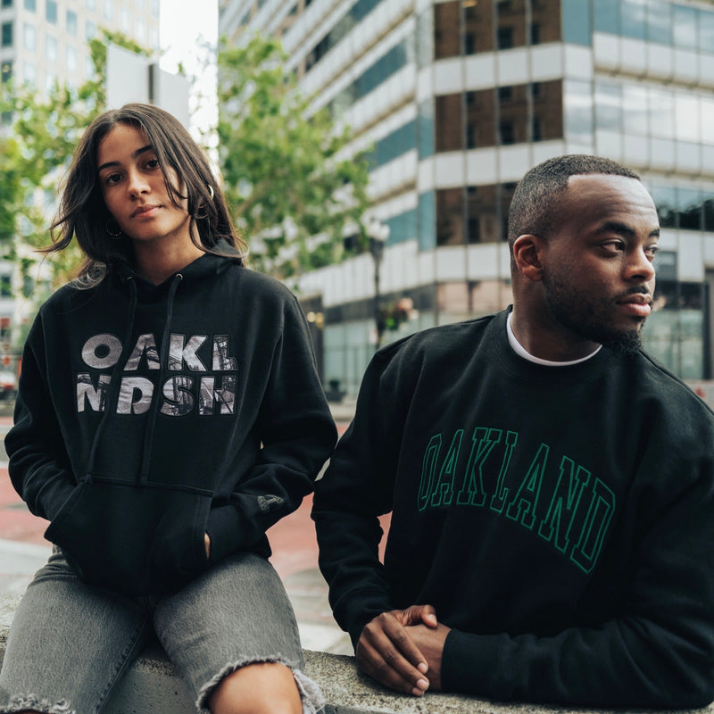A woman and a man sitting outdoors in Oakland wearing black Oaklandish sweatshirts.
