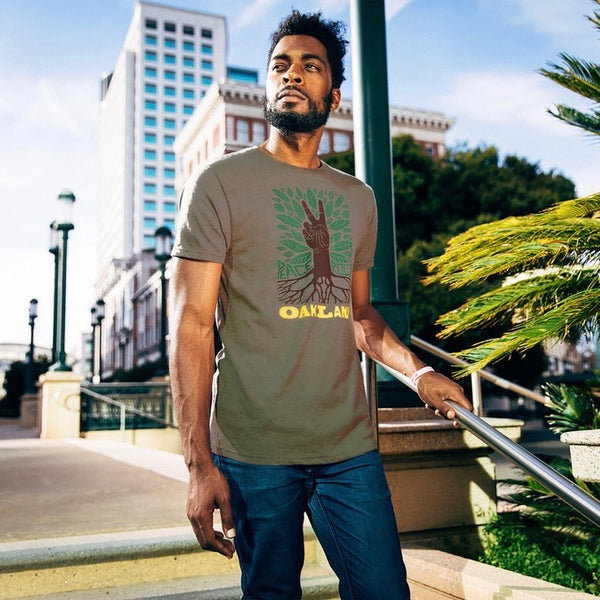 Army green t-shirt depicting an oak tree with green leaves with a brown hand for a trunk and words Peace, Roots, Oakland on male model.