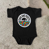 Black infant one-piece with Roots SC mosaic logo and wordmark on the chest lying on the asphalt.