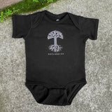 Black infant one-piece with a white Oaklandish tree logo and wordmark on the chest lying on the asphalt.