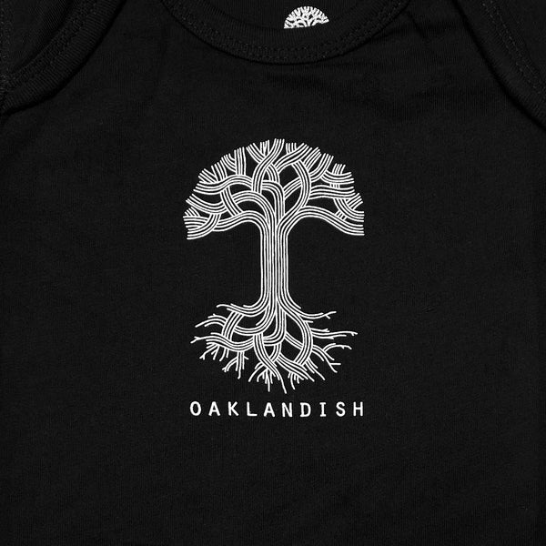 Close-up of the white Oaklandish tree logo and wordmark on a black infant one-piece chest.