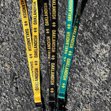 Three lanyards laying flat on black asphalt. One green, one black, and one yellow with Oaklandish wordmarks and tree logos.