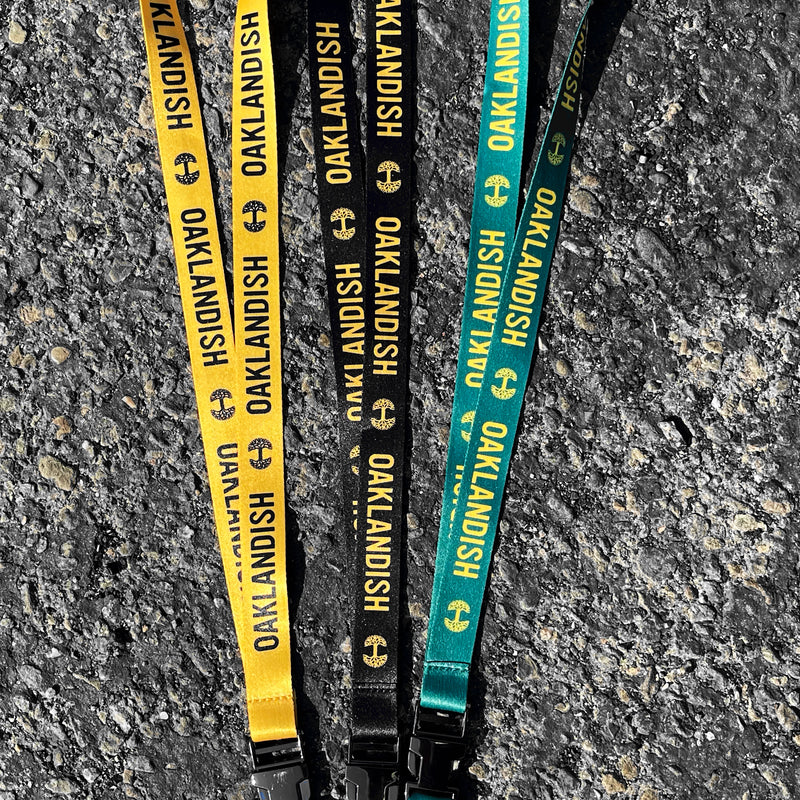 Three lanyards laying flat on black asphalt. One green, one black, and one yellow with Oaklandish wordmarks and tree logos.