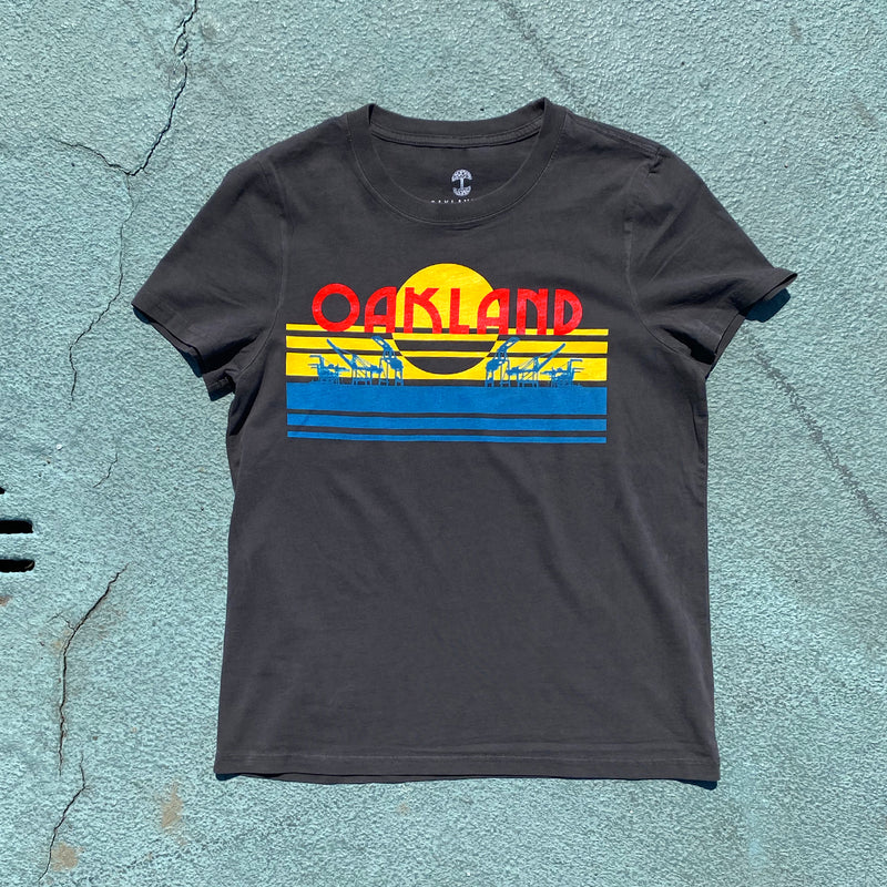 Faded black women’s t-shirt with multi-color license plate design with capital letters spelling out OAKLAND on blue asphalt.