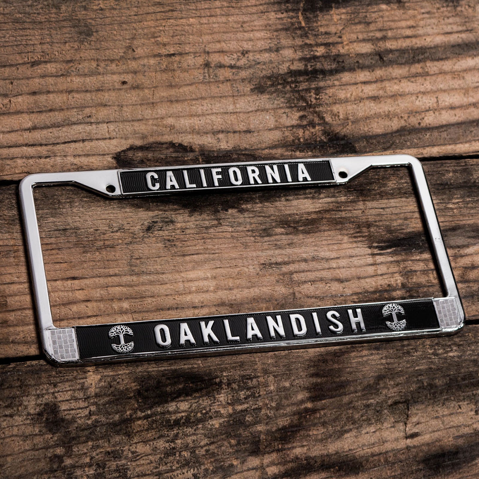 License plate holder with black rims with silver California wordmark on top and Oaklandish wordmark and tree logo on the bottom on wood desk.