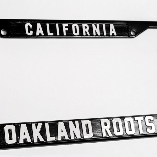 Close-up of California wordmark on top and Oakland Roots logo on the bottom of black rims on a silver license plate holder.