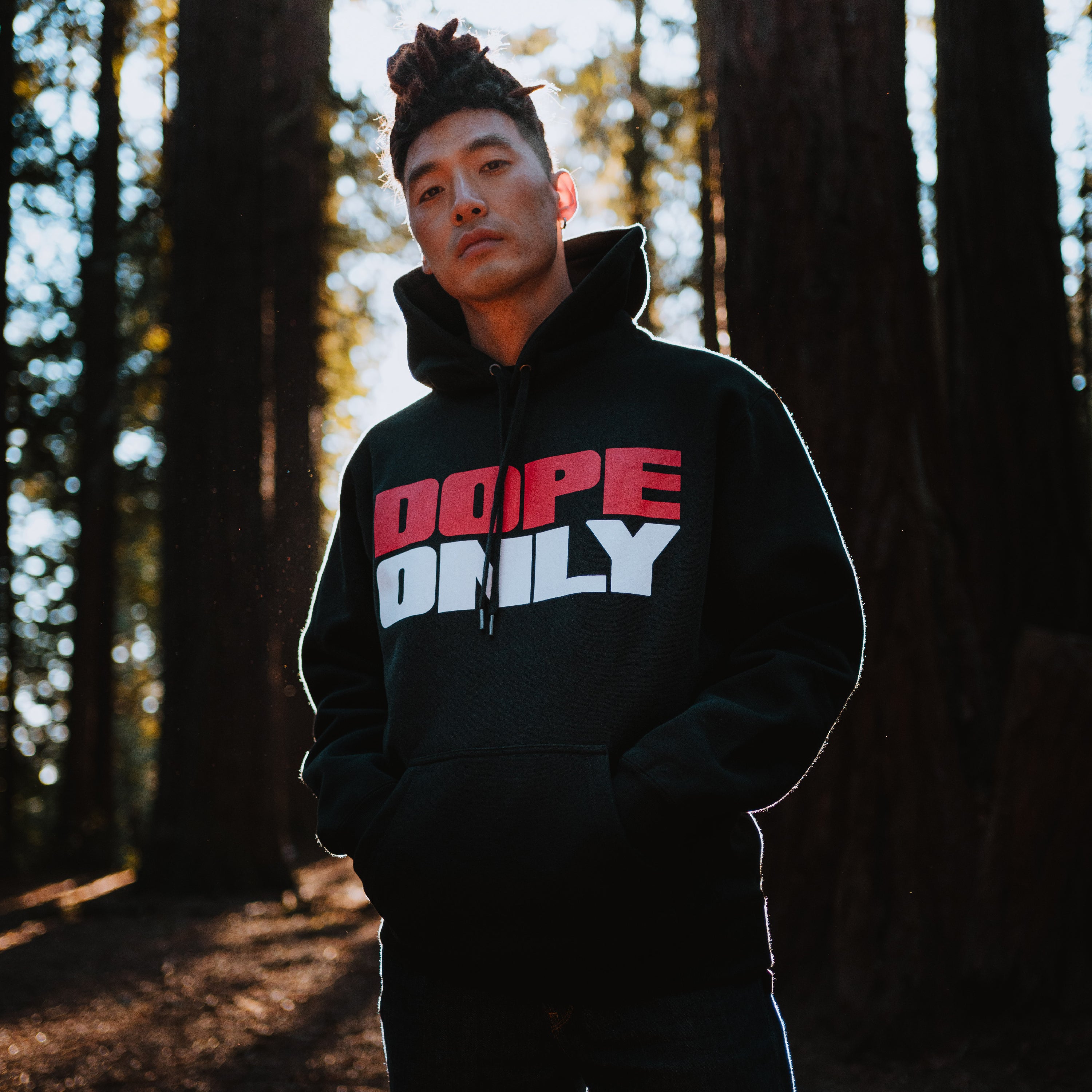 A man standing in a forest wearing a black hoodie with a red and white DOPEONLY wordmark logo on the chest.