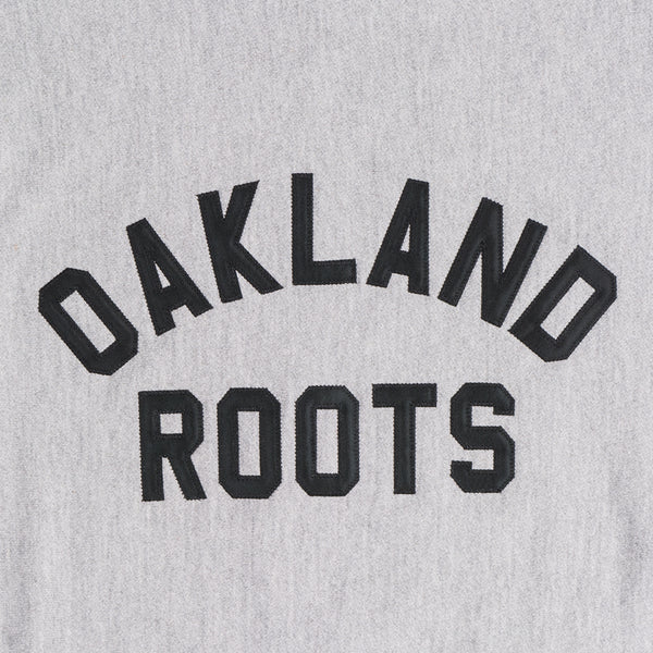 Close-up of black applique OAKLAND ROOTS wordmark on the chest of a grey crew sweatshirt.