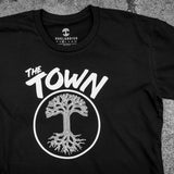 Close up of a black t-shirt with the words The Town and Oaklandish tree logo in a circle underneath on cement background.