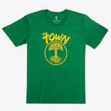 Green shirt flat on a plain background, shirt has a design in golden yellow ink saying the town and then the Oaklandish tree with roots logo inside of a circle.
