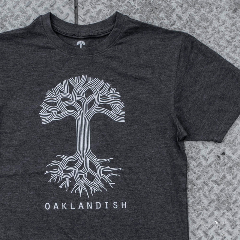 A charcoal heather t-shirt with a large white Oaklandish tree logo on the chest on a metal grate.