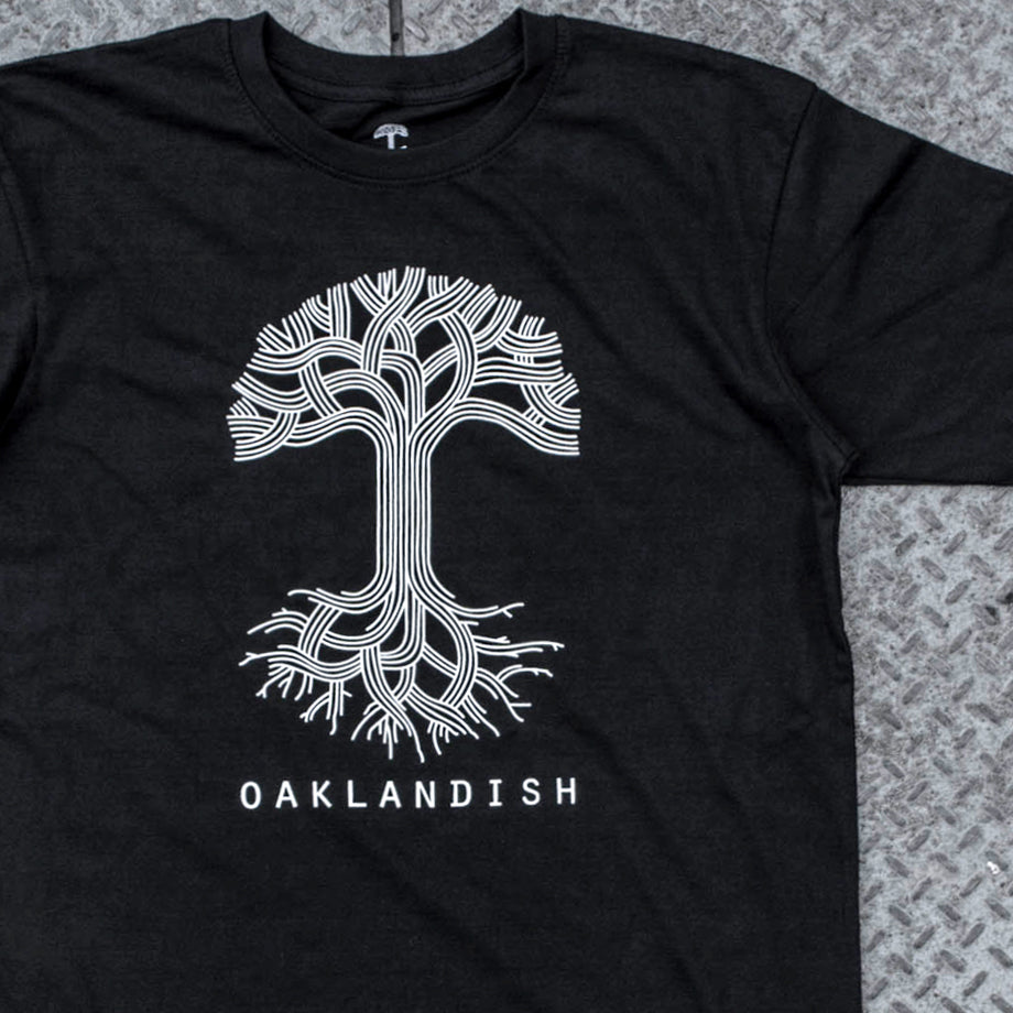 A black t-shirt with a large white Oaklandish tree logo on the chest is on a metal grate.