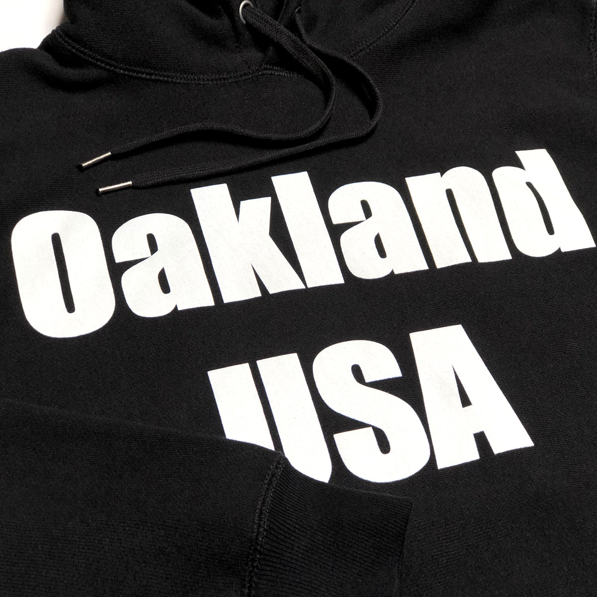 Close-up of large white Oakland USA wordmark logo on the chest of a black hoodie.